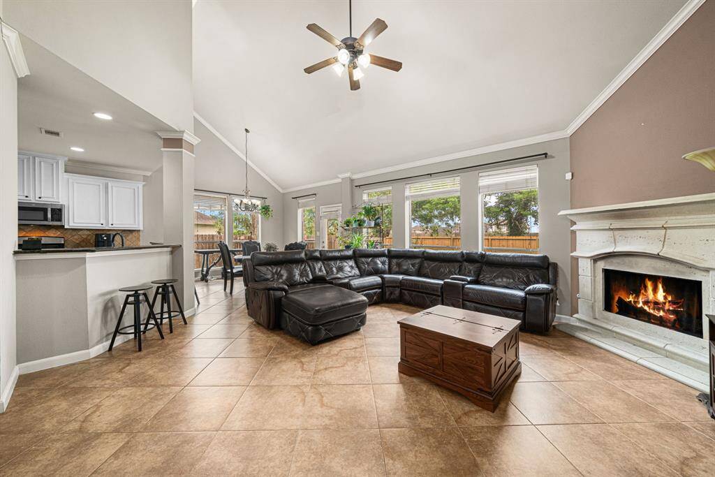 This expansive living area features soaring ceilings adorned with crown molding and a stunning stone fireplace, creating a warm and inviting focal point. The space is illuminated by oversized windows that frame picturesque backyard views while flooding the room with natural light. Practical elegance continues underfoot with durable tile flooring throughout. A convenient kitchen breakfast bar seamlessly connects this living space to the culinary area, perfect for both daily living and entertaining.