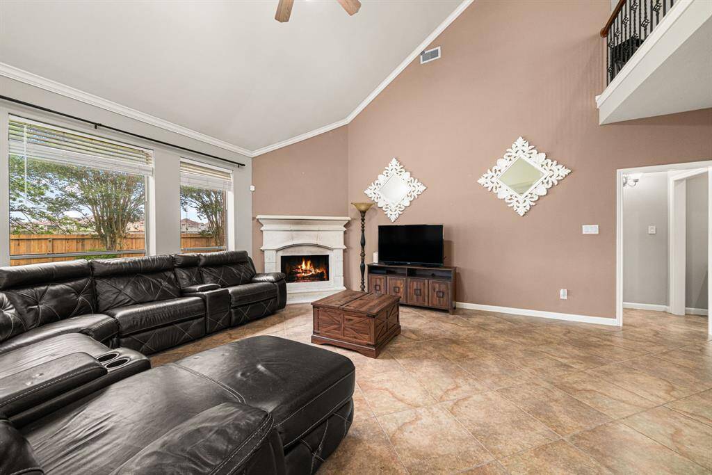 This expansive living area features soaring ceilings adorned with crown molding and a stunning stone fireplace, creating a warm and inviting focal point. The space is illuminated by oversized windows that frame picturesque backyard views while flooding the room with natural light. Practical elegance continues underfoot with durable tile flooring throughout. Second floor balcony overlooks the spacious living room.