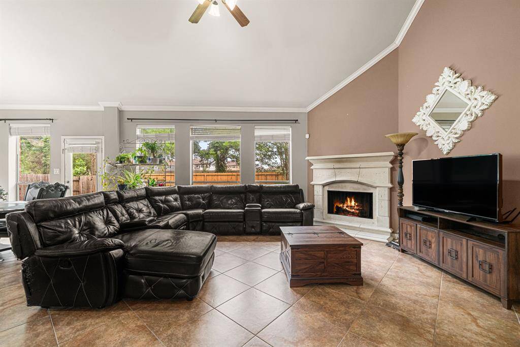This expansive living area features soaring ceilings adorned with crown molding and a stunning stone fireplace, creating a warm and inviting focal point. The space is illuminated by oversized windows that frame picturesque backyard views while flooding the room with natural light. Practical elegance continues underfoot with durable tile flooring throughout.