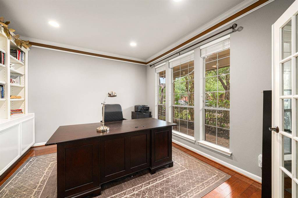 French doors open into a sophisticated study features three expansive windows framing picturesque front yard views, bathing the space in natural light. The room showcases custom built-in shelving, ample storage solutions, refined crown molding, and rich hardwood flooring - creating a perfect blend of functionality and timeless elegance.