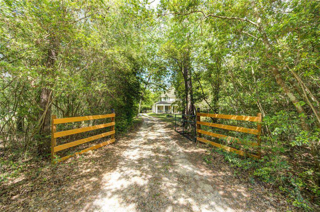 A charming property entrance with a gravel driveway leads through a wooded area, flanked by wooden gates. The path opens to a view of a cozy home nestled among lush greenery, providing a serene and private retreat.