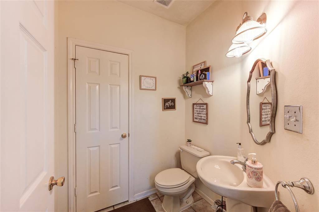 This is a small, cozy bathroom featuring a pedestal sink, a toilet, and a decorative mirror. The walls are adorned with shelves and framed art. It has soft lighting and neutral tones, creating a warm and inviting atmosphere.