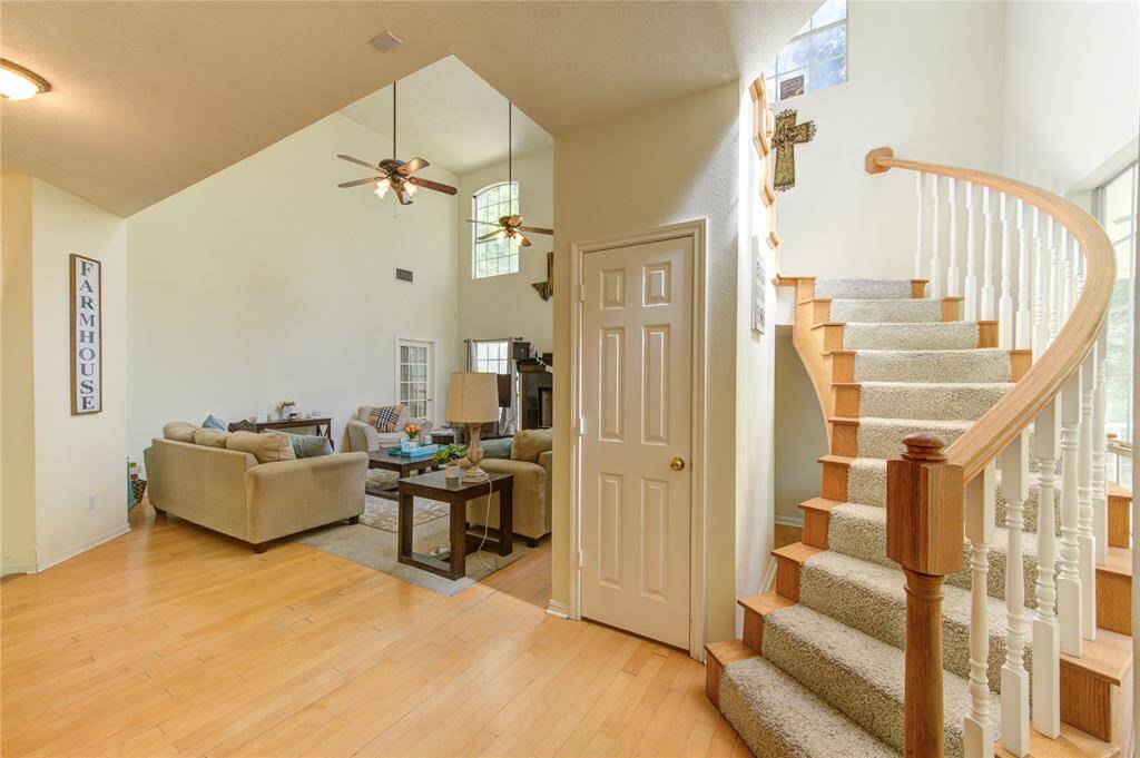 This inviting home features an open living area with high ceilings and large windows, allowing natural light to flow in. The space is enhanced by a cozy seating arrangement and a farmhouse decor accent. A curved staircase with carpeted steps and wooden railing adds elegance, leading to the upper level. The light wood flooring complements the neutral tones of the furnishings.