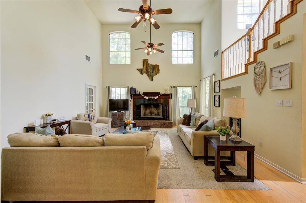 This spacious living room features high ceilings and large windows, allowing plenty of natural light. It includes a cozy fireplace, elegant staircase, and is tastefully furnished with neutral tones, creating a welcoming atmosphere.