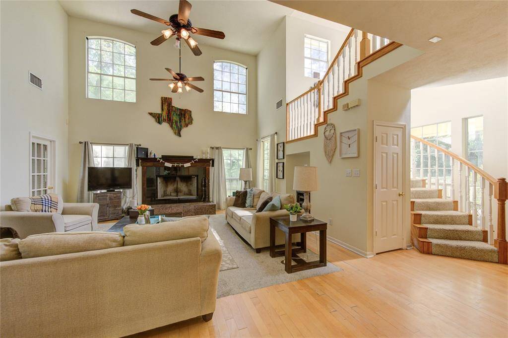 This inviting living room features a high ceiling and abundant natural light from large windows. It has a cozy fireplace, modern ceiling fans, and an open staircase, creating a warm and spacious atmosphere.