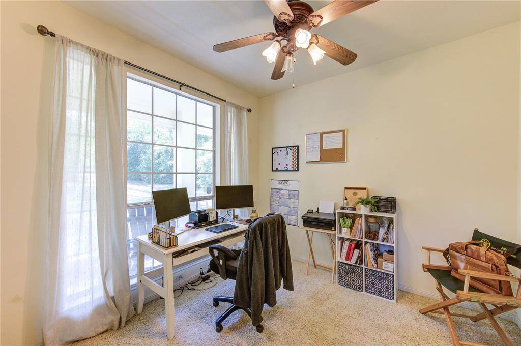 Bright home office with large window, ceiling fan, and carpeted floor. Features a tidy desk setup and shelving for organization.