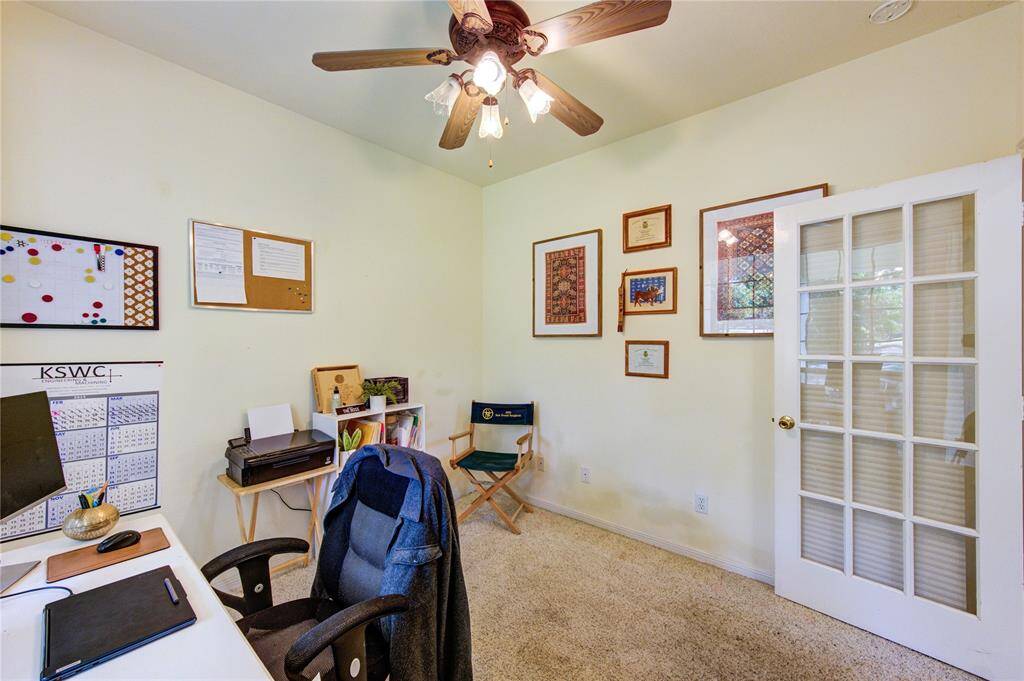 This cozy home office features a ceiling fan, carpeted flooring, and a glass French door. It includes a desk, chair, and wall-mounted decorations, creating a functional and inviting workspace.