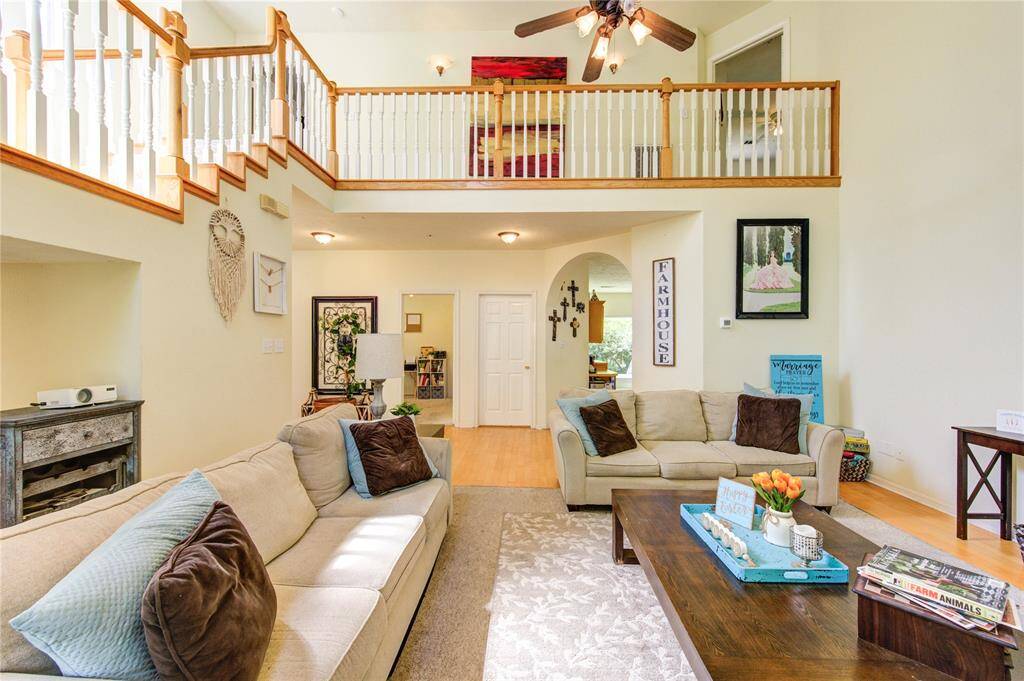 This spacious living room features high ceilings and abundant natural light. There is a cozy seating area with plush sofas, a wooden coffee table, and tasteful decor. A balcony overlooks the room, adding an open and airy feel, while light wood flooring complements the neutral walls.