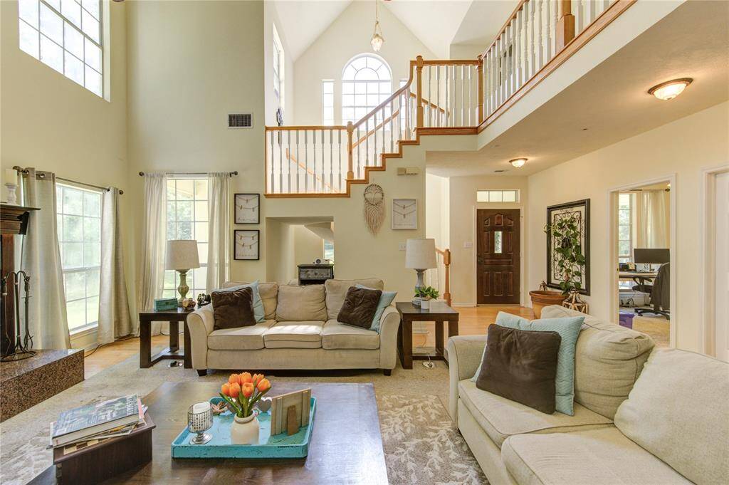 This spacious living room features high ceilings and large windows, creating a bright and airy atmosphere. It includes a cozy seating area with a fireplace, and an open staircase leading to the upper level, adding a touch of elegance.