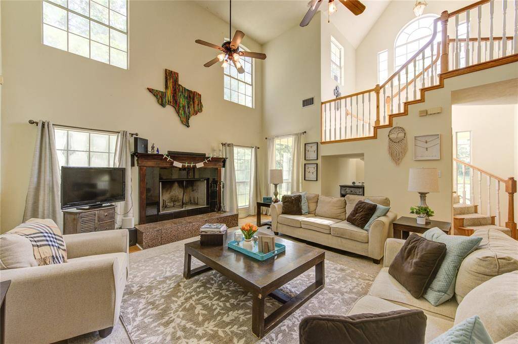 This spacious living room features high ceilings and abundant natural light. It includes a cozy fireplace, modern ceiling fans, and an open staircase leading to the upper level. The room is tastefully decorated with neutral tones, providing a warm and inviting atmosphere.