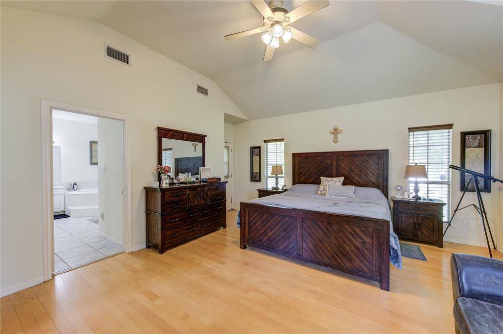 This spacious bedroom features a vaulted ceiling, wooden floors, and ample natural light from two windows. It includes a large bed, matching furniture, and an ensuite bathroom with a bathtub visible through an open door. A ceiling fan provides added comfort.