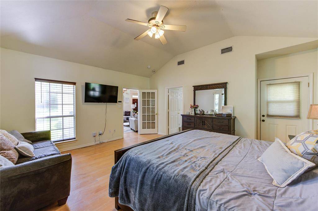 This spacious bedroom features a vaulted ceiling with a ceiling fan, natural light from large windows, and a cozy sitting area. It includes a wall-mounted TV, a dresser, and a door leading to another room, offering convenience and comfort.