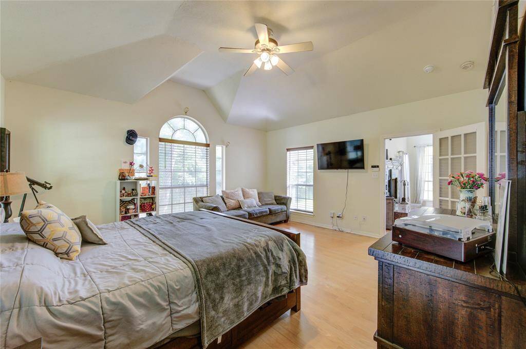 Spacious bedroom with vaulted ceiling, large arched window, and ample natural light. Features include a ceiling fan, wall-mounted TV, and cozy seating area. The room has light wood flooring and connects to another room via French doors.