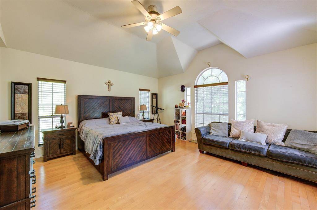 Spacious bedroom with vaulted ceiling, featuring a large bed, wood flooring, and ample natural light from multiple windows. Cozy seating area with a plush sofa complements the room.