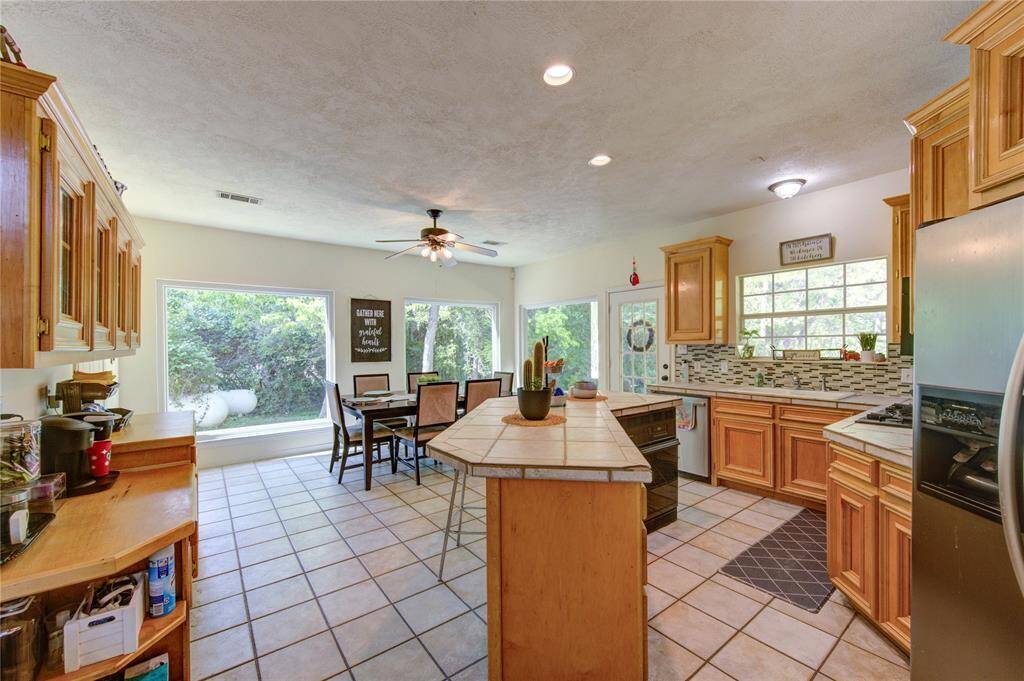 This kitchen features warm wood cabinetry, a central island, and stainless steel appliances. Large windows provide natural light and a view of the greenery outside. There's a cozy dining area with a ceiling fan, perfect for family meals. The space is open and inviting.