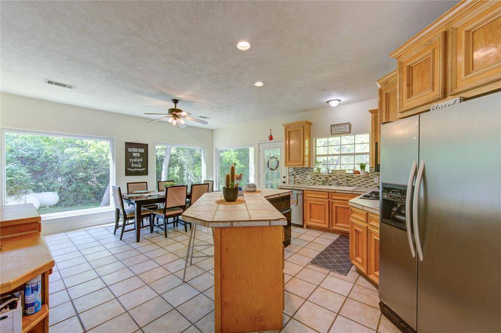 Bright and spacious kitchen with large windows showcasing greenery, featuring a dining area, modern stainless steel appliances, ample wooden cabinetry, and a central island.