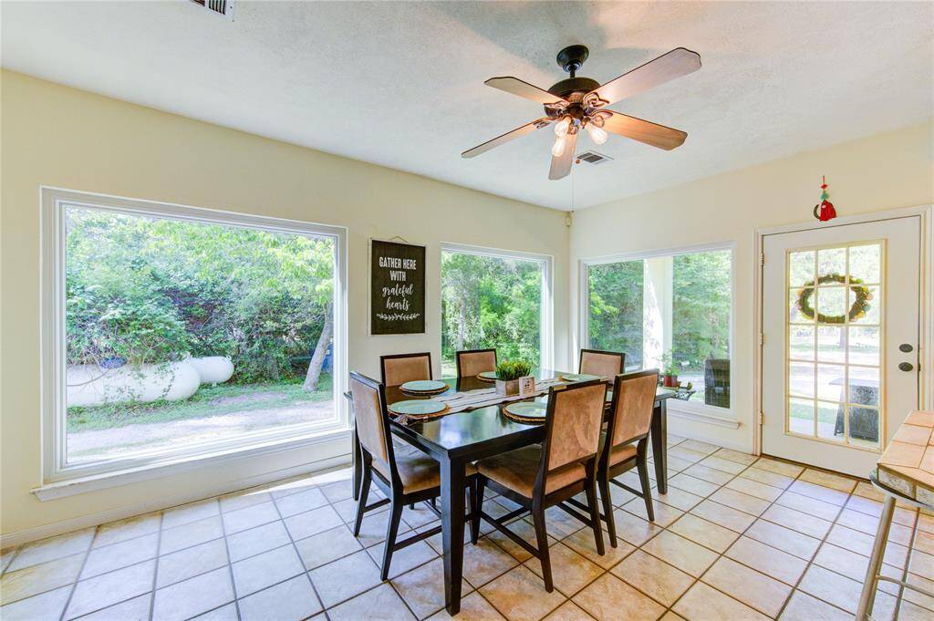 Bright dining area with large windows, tile floors, and a ceiling fan. The space includes a table for six and offers a view of greenery outside.