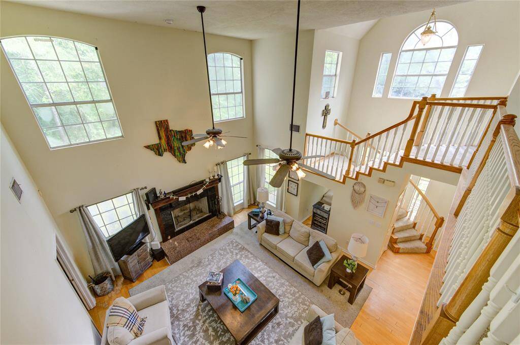 This spacious living room features high ceilings and abundant natural light from large windows. A fireplace serves as a cozy focal point, while an open staircase and loft area enhance the airy feel. The layout is ideal for both relaxation and entertaining.