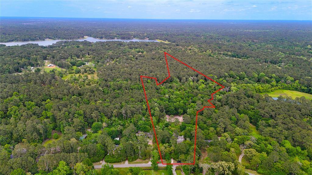 This aerial photo shows a large, wooded property outlined in red, nestled in a lush, green landscape. The area is expansive and surrounded by dense forest, offering privacy and a serene environment. Nearby are a few scattered homes and a distant view of a lake, providing a sense of rural tranquility.