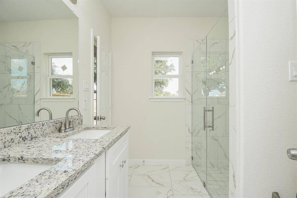 Modern bathroom with marble tiles, a glass shower door, granite countertop, and two windows allowing for natural light.