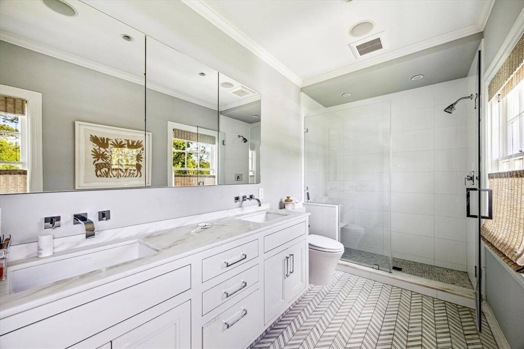 Effortlessly modern, the primary bathroom features a double vanity with marble countertops, sleek wall-mounted fixtures, and a spacious glass-enclosed shower. Chevron-patterned tile flooring adds a stylish, contemporary touch to this serene retreat.