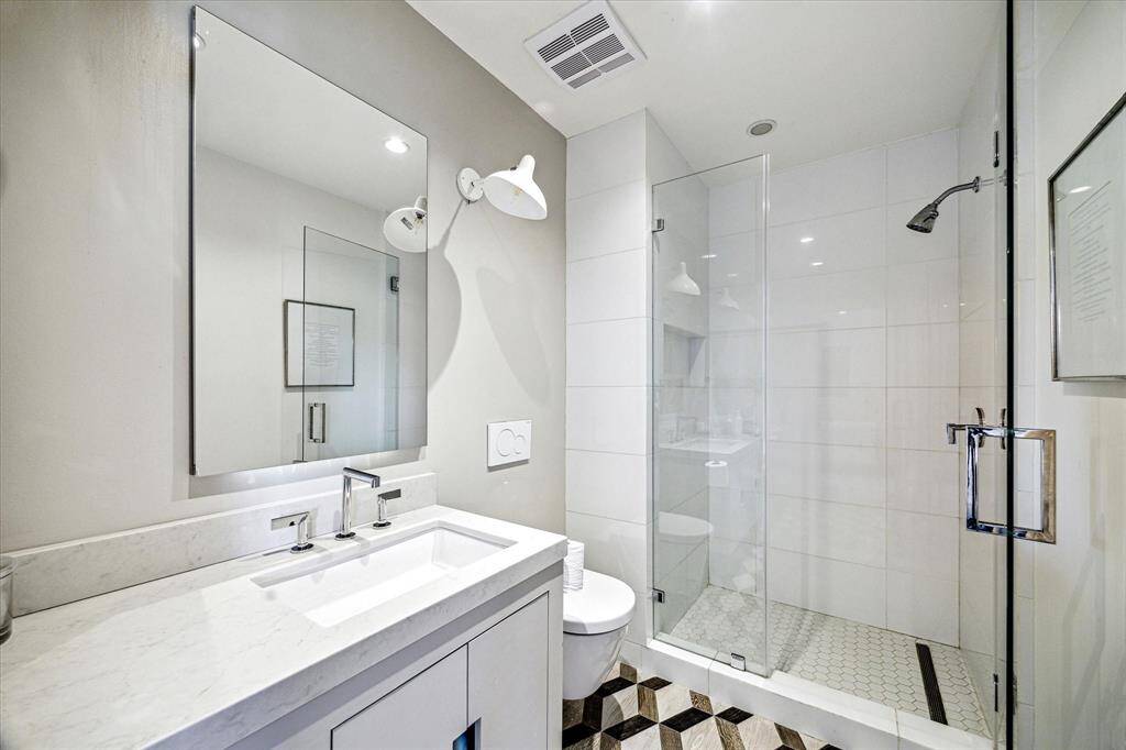 Stylish and streamlined, this secondary bathroom features a large vanity, custom built-in cabinetry, oversized mirror, and glass-enclosed shower with penny tile flooring. Bold geometric tile underfoot adds a touch of personality to the crisp, modern design.