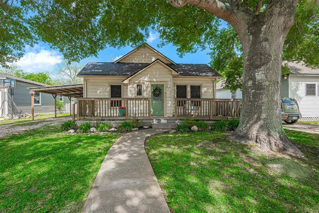 Charming single-story home with a welcoming front porch and a spacious yard. Features a large tree providing shade and a paved walkway leading to a distinctive front door. Ideal for cozy living with great curb appeal.