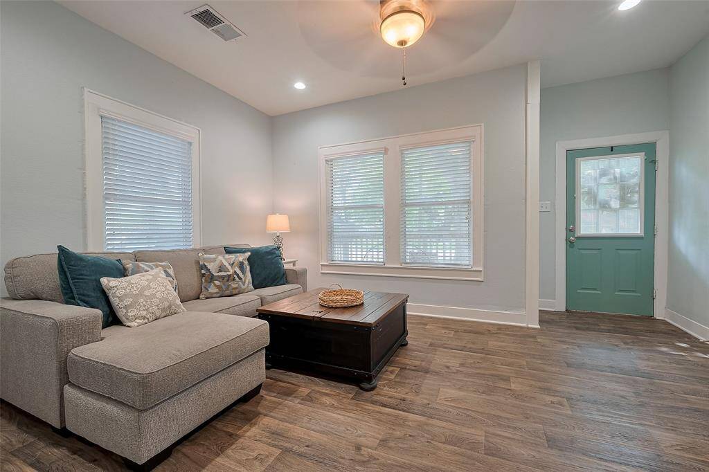 This cozy living room blends comfort and charm with wood-look flooring, soft, neutral toned blue walls, and large windows that fill the space with natural light. A ceiling fan adds a breezy touch, perfect for laid-back country living.