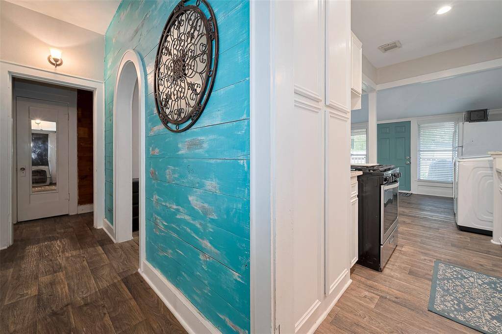 This photo showcases a charming hallway featuring rustic wood-look flooring and a striking turquoise accent wall. The space leads into a bright kitchen area with modern appliances and ample natural light from large windows.