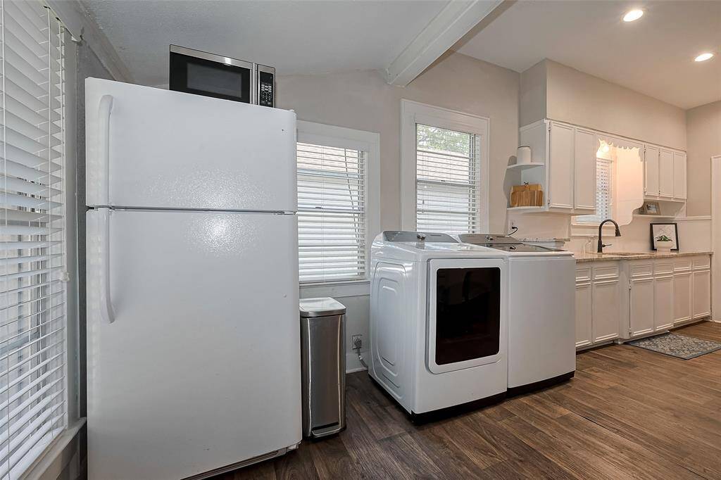 This photo showcases a bright, functional utility area featuring a full-size refrigerator, modern washer and dryer, and ample cabinetry for storage. The room has large windows for natural light and wood-look flooring, creating a clean, inviting space.