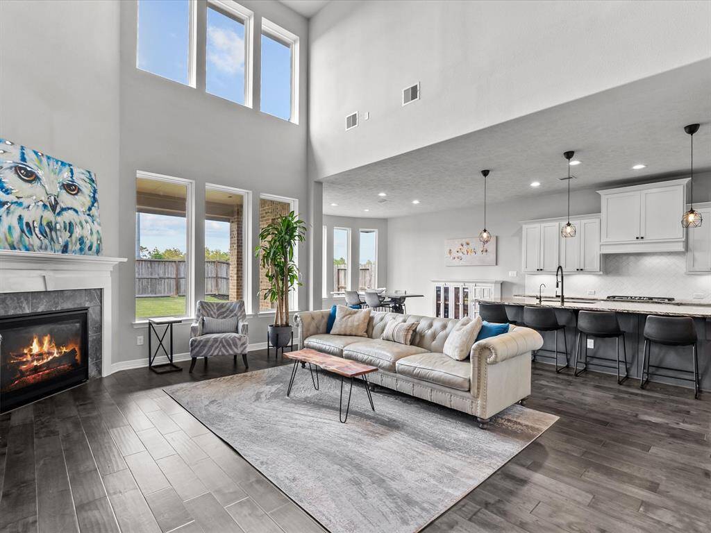 The living room open to the kitchen features wood flooring, two-story high ceilings, a wall of windows and a cozy fireplace for those cold winter nights