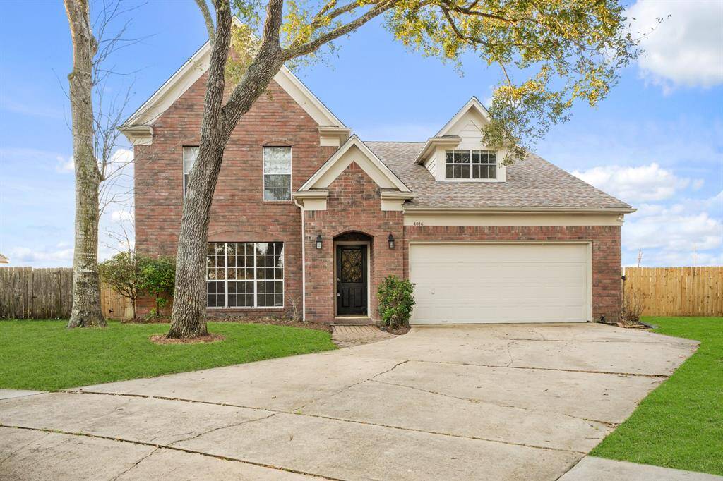 Charming two-story brick house with a gable roof and a large two-car garage. The front yard features a spacious driveway and mature trees, along with a well-maintained lawn. The home has a welcoming entrance with a decorative front door and large windows that allow for plenty of natural light.