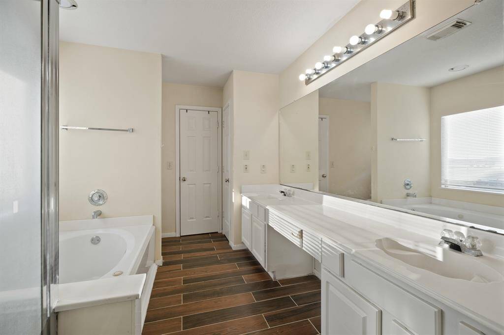 Spacious primary bathroom featuring a large vanity with dual sinks, a wide mirror with plenty of lighting, a bathtub, and a separate shower. The room is bright with natural light from a window, and has a stylish wood-look tile floor.