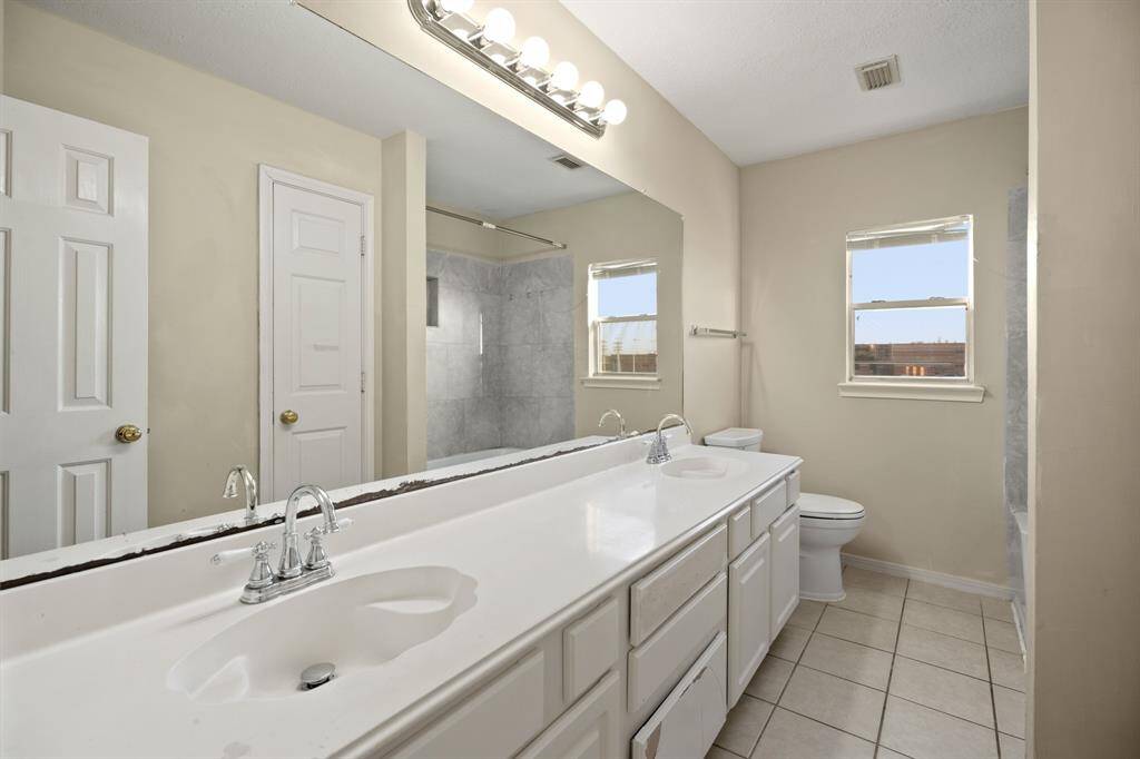 This is a spacious alternatebathroom featuring a long double-sink vanity with ample storage. It has a large mirror, tiled flooring, and a shower-tub combination. The room is well-lit with natural light from the window.