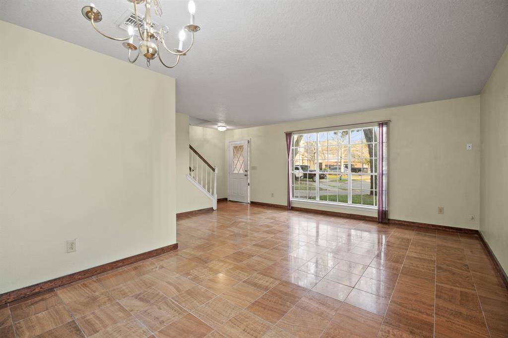 This spacious, light-filled living area features a large window overlooking the front yard, tiled flooring, and a classic chandelier. The room leads to a staircase and front entrance, offering a welcoming and open layout.