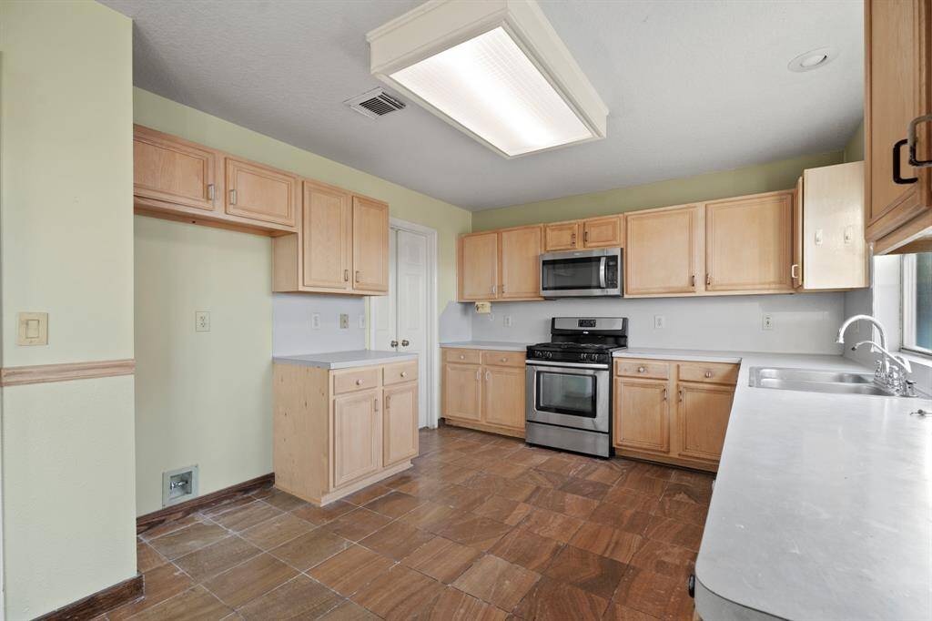 This kitchen features light wood cabinetry, a stainless steel gas stove and microwave, and ample counter space. It has a spacious layout with tile flooring.
