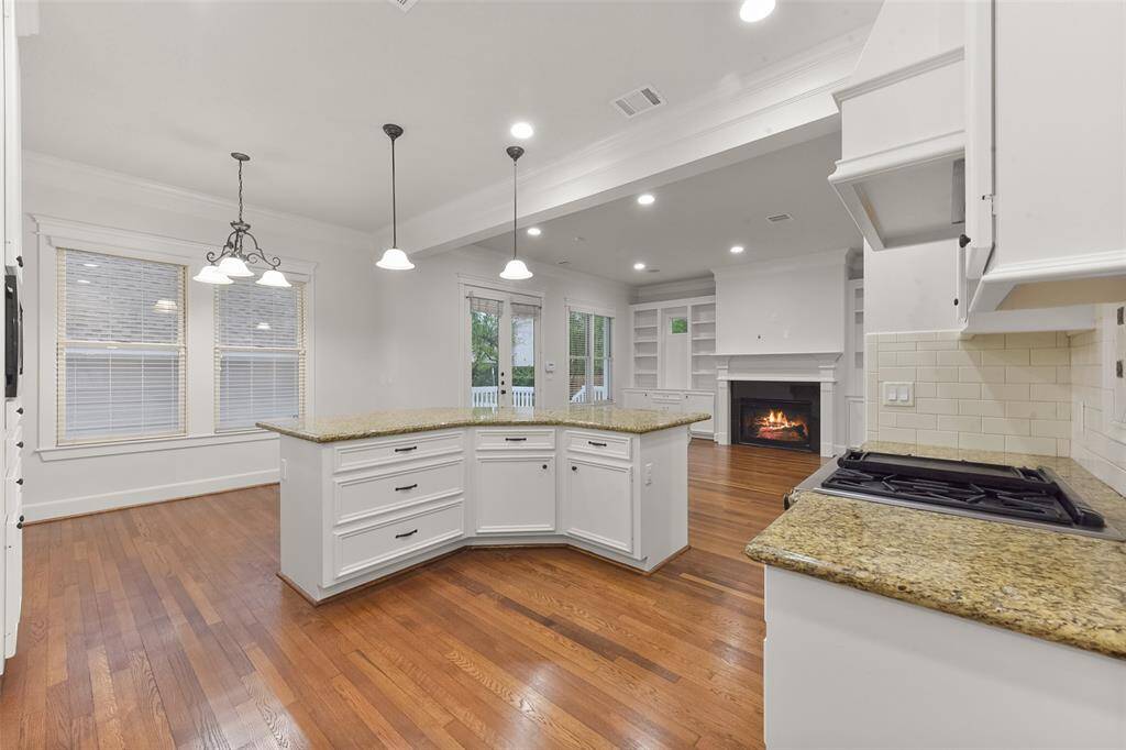 A spacious, open-concept kitchen and living area with hardwood floors. The kitchen features granite countertops, white cabinetry, and modern lighting. The family room includes built-in custom cabinets and a cozy fireplace, creating an inviting atmosphere. Large windows provide ample natural light and a view of the outdoor space.