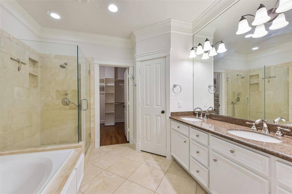 This primary bathroom features a spacious layout with a double vanity, ample lighting, and a large mirror. It includes a glass-enclosed shower, a separate jetted bathtub, and access to a large walk-in closet. The neutral tile flooring and walls create a clean, modern look.