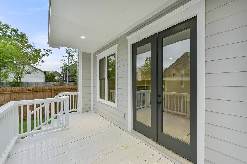 A charming outdoor back porch with light gray siding and double glass door leads to a fenced backyard. The space is well-lit and features a white railing, offering a cozy spot for relaxation.