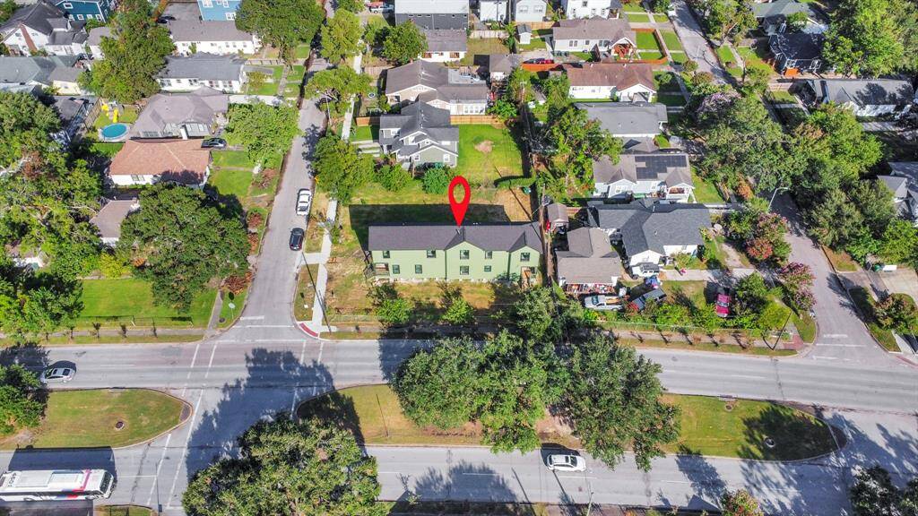The photo shows an aerial view of a residential neighborhood with a two-story house marked by a red arrow. The property is surrounded by lush greenery and is located at the intersection of two streets, offering easy access and potentially more visibility. The neighborhood appears well-established with a mix of home styles and mature trees.