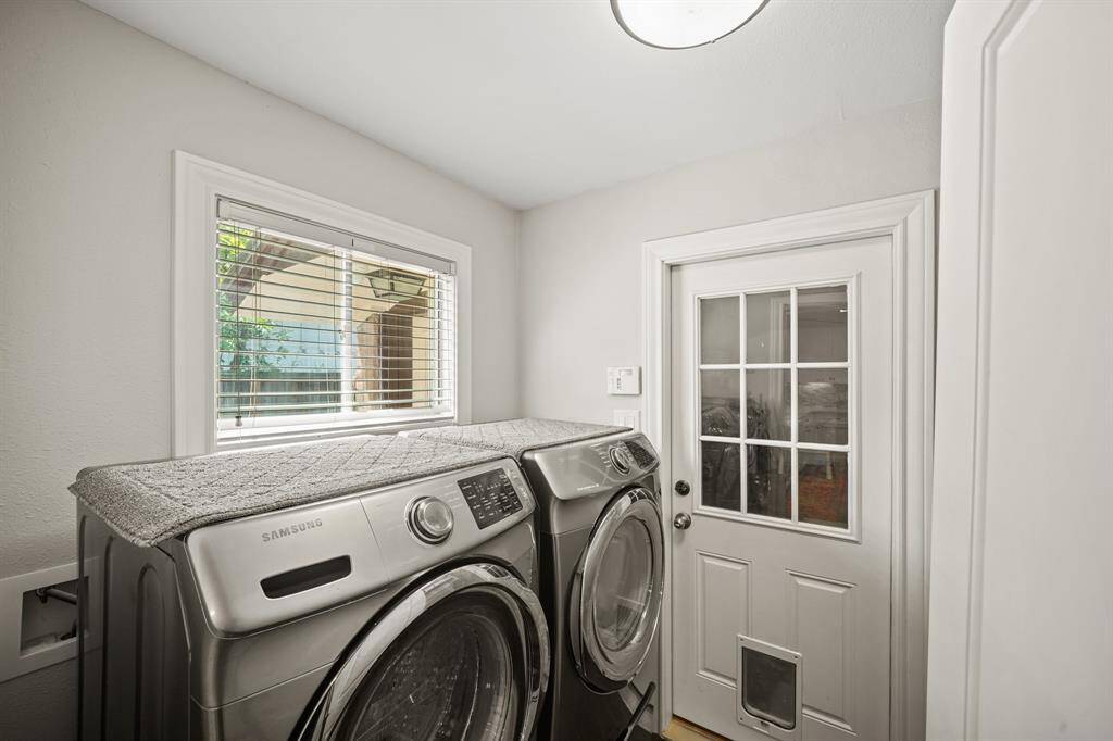 The Laundry Room offers Walnut Hued Handscraped Hardwood Floors, Full Size Samsung Front Loading Washer and Dryer (with Elevated Stand and Drawers) remains per Seller, Wall Mounted Shelf, Hanging Rod, Faux Wood Blinds, Light Grey Designer Paint, Flush Mount Light Fixture, Raised Water Hater on a Platform, and Extended Baseboards.