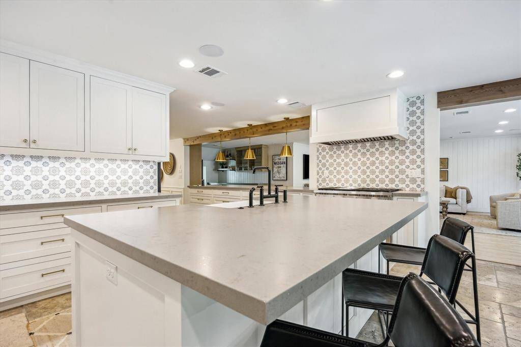 This modern kitchen features a large island with a sleek countertop, marble backsplash, and ample white cabinetry. It includes black bar stools, recessed lighting, and a cozy view into the living area, creating an inviting open-concept space.