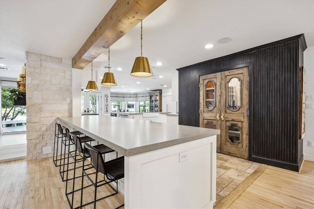 This modern kitchen boasts a spacious pensilua with seating, pendant lighting, and a blend of sleek and rustic elements. It features a stone accent wall, wooden ceiling beam, and ornate double doors leading to a French pantry, creating a welcoming and stylish atmosphere.
