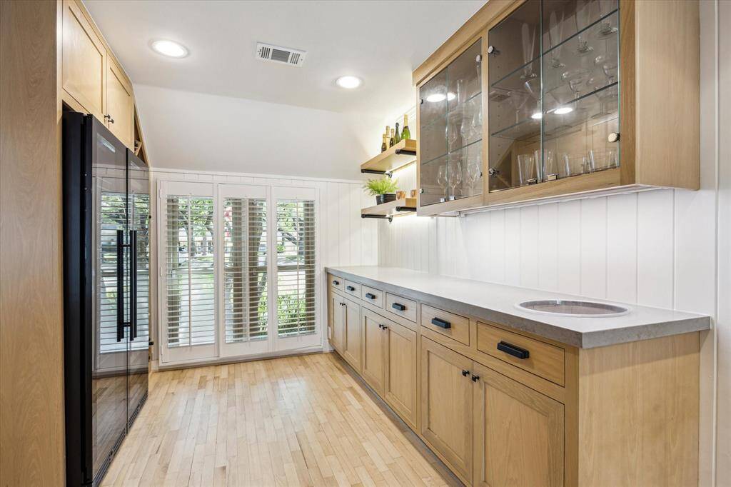 This spacious and bright butler's pantry features light wood cabinetry, glass-front upper cabinets, and open shelving for ample storage. It includes a modern wine fridge (2025), beverage fridge (2025), sleek countertop, and a small ice bucket, with large windows and shutters providing natural light and a view. Perfect for entertaining and additional kitchen storage.