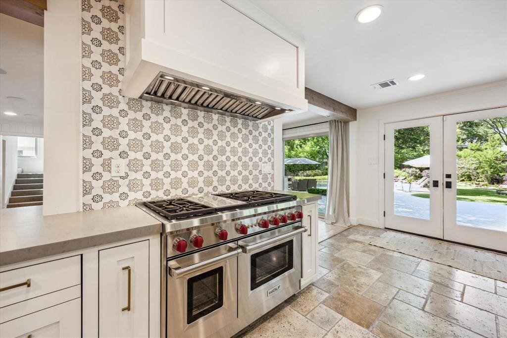 This kitchen features a marble backsplash, Wolf gas range with 6 burners, a griddle and double ovens, and light cabinetry. French doors open to a lush backyard, providing ample natural light and a seamless indoor-outdoor transition.