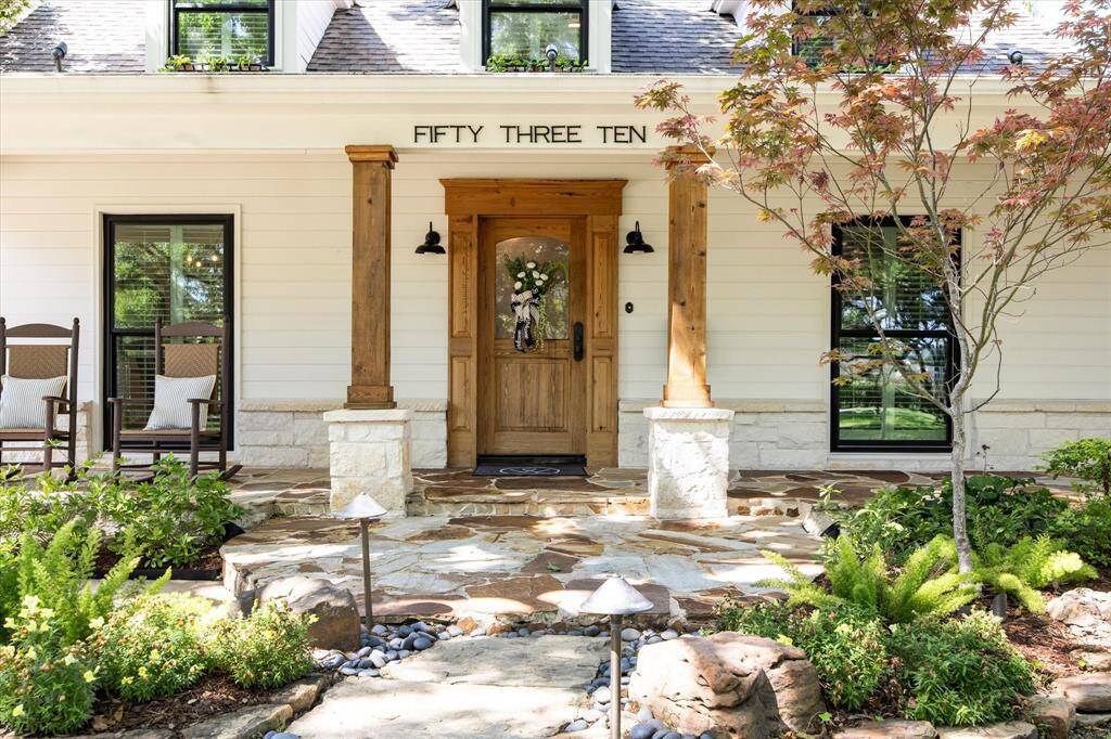 This charming home's entrance features a welcoming wooden door framed by stone pillars, a well-maintained garden with lush greenery, and a cozy porch with rocking chairs. Welcome home!
