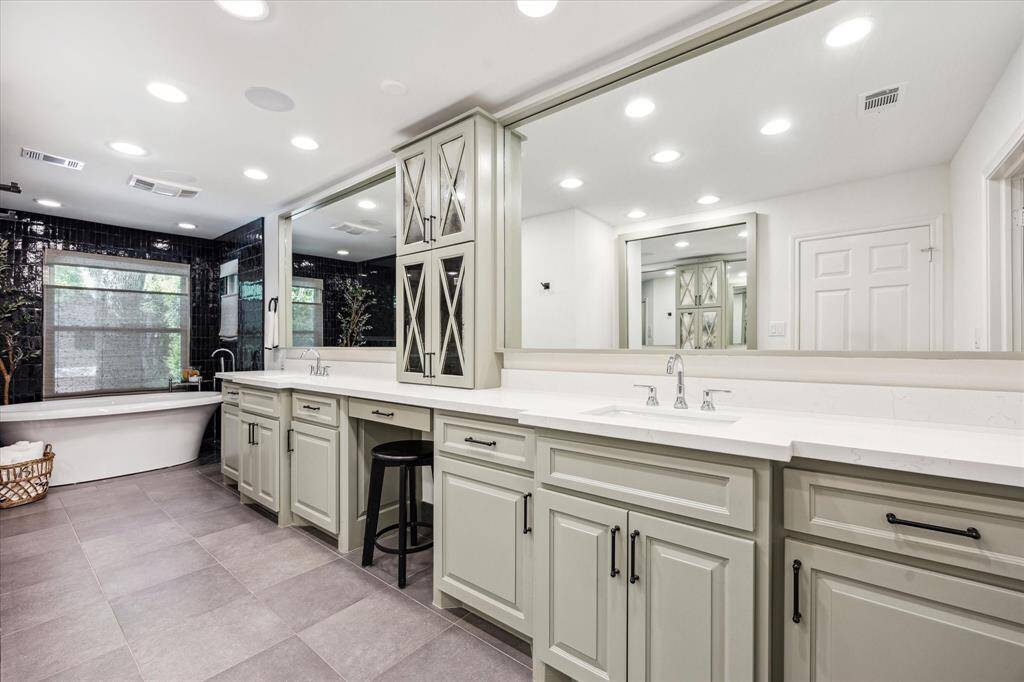 This spacious primary bathroom features dual sinks with ample counter space, elegant cabinetry, a vanity with make-up station, a large mirror, and a freestanding bathtub. The room is well-lit with recessed lighting and has modern tile flooring, creating a luxurious and functional space.