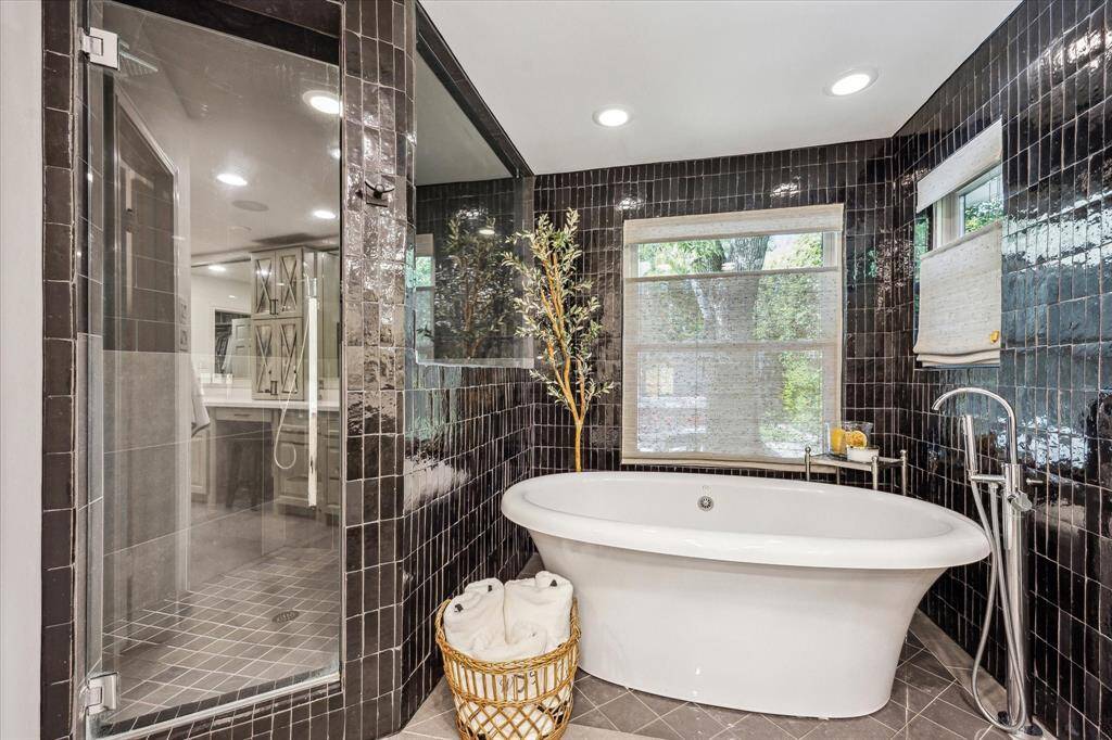 The primary bathroom features a sleek design with glossy black tiles and a large window providing natural light. It includes a modern freestanding bathtub, a spacious glass-enclosed shower, and elegant fixtures, creating a luxurious and tranquil atmosphere.