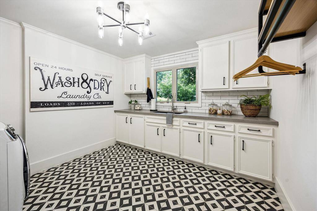 This spacious laundry room features stylish black and white patterned flooring, a beautiful custom light fixture, ample white cabinetry, and a large window for natural light.