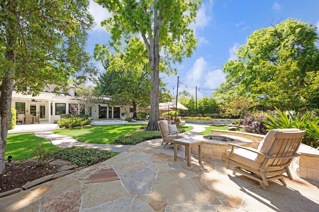 This photo showcases a spacious, inviting backyard with a large patio area, lush greenery, and mature trees. The patio features a stone fire pit and comfortable seating, ideal for outdoor relaxation and entertaining. The well-maintained garden and string lights add charm and ambiance.